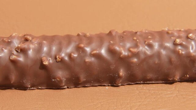 Slow orbiting close-up around a chocolate coated snack bar with crunchy nut pieces on a warm brown studio background, showing glossy texture and product detail.