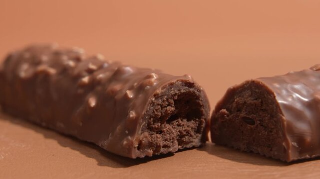 Slow tracking shot to the left across cut chocolate coated snack bars and a whole bar, showing textured filling, glossy coating and nut pieces in a warm studio setting.
