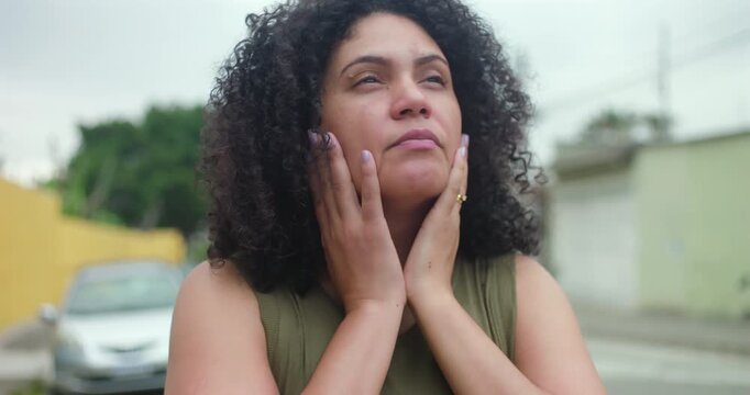 Woman standing outdoors with hand on face, looking down in deep thought, emotional distress, contemplation, concern, and personal struggle in everyday urban environment