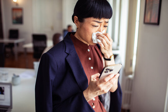 Businesswoman blowing nose while reading smartphone in office