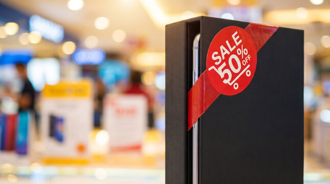 Smartphone retail box with red sale seal in a blurred electronics store. Black Friday product promotion for holiday shopping, technology sales, and retail advertising