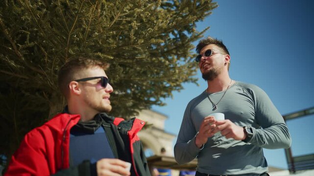 Two male friends enjoy a casual conversation outdoors on a sunny winter day. They share a relaxed moment with warm drinks.