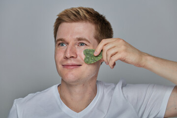 A young man gently massages under his eye with a jade gua sha stone during a calming skincare routine, highlighting self-care, facial massage and natural wellness for glowing skin. © Kateryna Zghurska