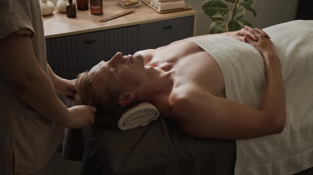 Therapist performing head and neck massage on relaxed Caucasian young adult man lying on spa table. Ideal visual for wellness marketing, luxury resort promotion, and physical therapy advertising