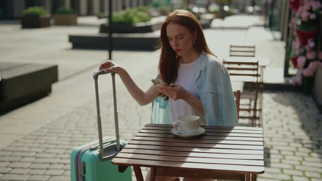 Anxious young woman sitting at outdoor cafe with luggage, looking for taxi checking phone, getting frustrated and worried about being late for trip, checking time and feeling annoyed, slow motion.