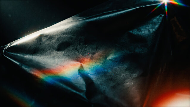 Crumpled dark plastic foil texture with iridescent rainbow prism light reflection and anamorphic lens flare