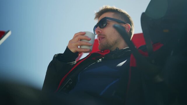A man in sunglasses sips from a mug, enjoying a moment of relaxation outdoors. He takes a break, drinking a warm beverage under the clear blue sky.