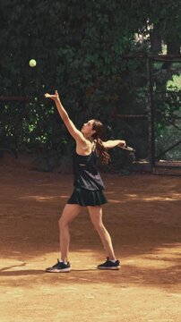 Competitive woman serves tennis ball and continues energetic rally on outdoor clay court with focused determination. Tennis match, sports training, active fitness concept.