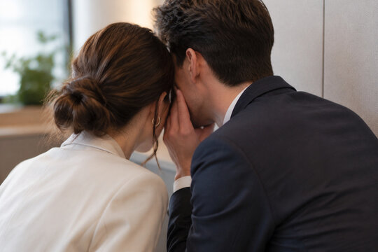 Two adults leaning close in office corner whispering, workplace gossip and secrecy concept
