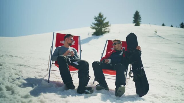 Two friends relax in deck chairs on a sunny, snowy mountain with a snowboard. They enjoy a warm drink during their winter sports break.