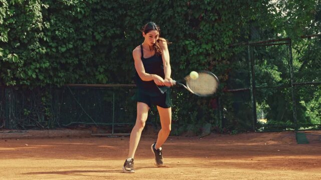 Determined female tennis player delivers powerful serve and continues rally on outdoor clay court with competitive energy. Tennis competition, athletic training, sports performance concept.