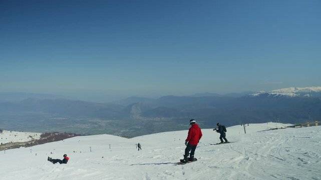 A snowboarder and skiers enjoy a clear day on snow-covered mountain slopes. The scene depicts active recreation amidst a breathtaking winter landscape.