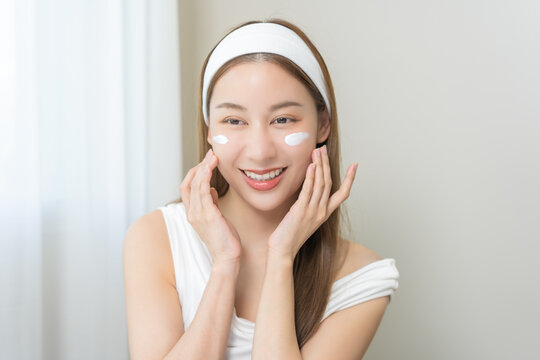 Facial beauty, skin care concept, smile asian young woman applying moisturizer lotion on her face, pretty girl putting cream treatment before makeup cosmetic. Isolated on background.