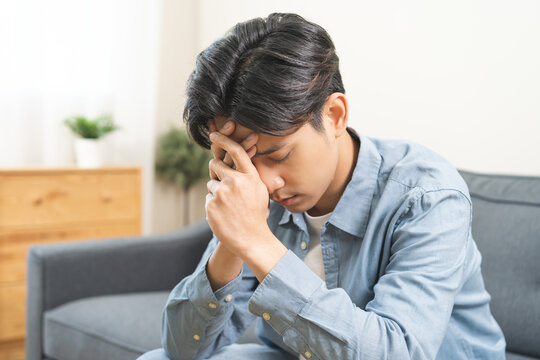 Sad unhappy, alone asian young man headache holding forehead thinking about problem, difficulty, feeling failure and exhausted, suffering sitting on sofa, grief sorrow and bad relationship or break up