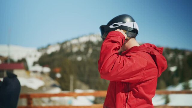 A man in a red jacket fastens his ski helmet and goggles. He is ready for winter sports in the snowy mountains.