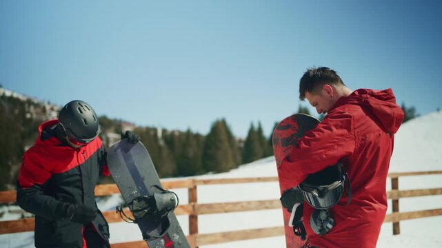 Two male snowboarders in red jackets adjust their snowboards and gear on a sunny winter mountain. They are preparing for a thrilling day of winter sports and adventure on the snowy slopes.