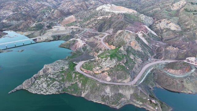High aerial flight crossing a bridge from a construction site toward a sand quarry, with heavy machinery and a road visible beside the lake.