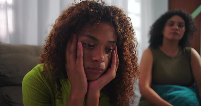 Teenage daughter feeling sad, sitting on couch with hands on face, mother in background reaching out, showing concern and trying to comfort her