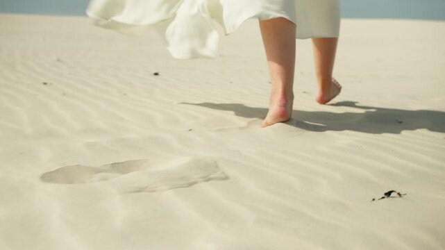 Woman's bare feet walking on sand dunes leaving footprints as her white dress flutters in the wind on a sunny beach day