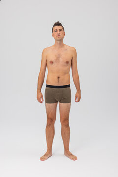 Full-body front view portrait of a bare-chested man with a hairy chest and legs, standing in olive green boxer shorts against a light grey studio background