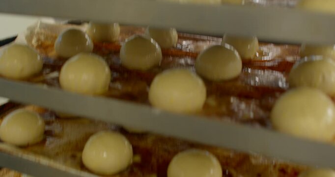 low-angle close-up of round glossy dough portions on parchment-lined metal trays stacked in bakery rack, warm light reflections, dough texture for proofing food