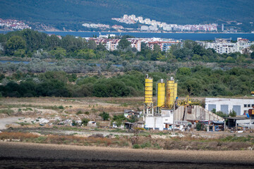 An industrial concrete batching plant featuring tall yellow cement silos and aggregate loading ramps sits in an arid valley, framed by lush tree lines © Andrey