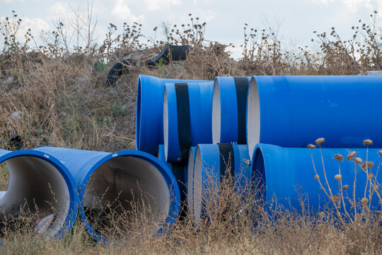 Large-diameter blue water pipes with pale interior linings sit stockpiled on dry, weed-covered ground near an earthen embankment at an outdoor construction staging site.