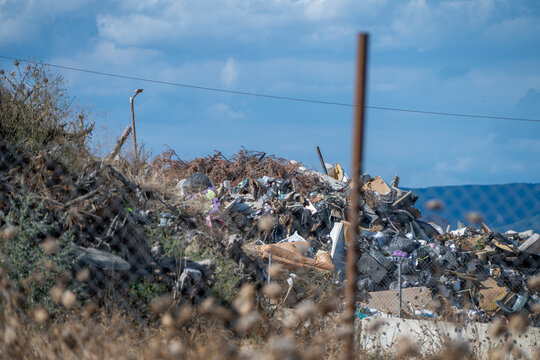 A sprawling mound of solid municipal waste and scrap debris sits behind an old wire fence post in an outdoor landfill depot beneath a cloudy sky.