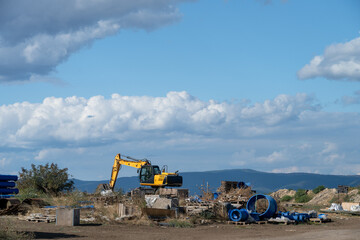 A yellow tracked excavator sits on an open construction supply staging ground scattered with pallets, industrial piping, and earthwork mounds against distant mountain foothills and a cloudy sky. © Andrey