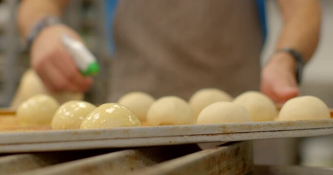 close-up low-angle shot of baker applying liquid glaze from yellow spray bottle with green nozzle onto evenly spaced round dough balls on metal baking tray