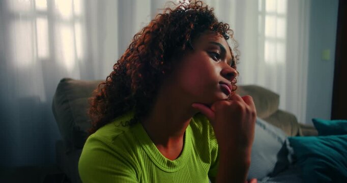 Young African descent girl looking outside, resting her chin on her hand, thoughtful moment in a cozy living room with natural light and calm atmosphere