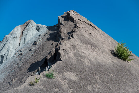 A steep, eroded mound of fine gravel and quarry slag rises sharply into a vibrant blue sky, dotted with a solitary green shrub clinging to its slope.