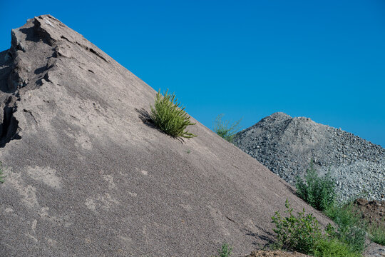 A sharply ridged, steep mound of excavated gravel and slag rises into a vivid blue sky, with a lone green shrub clinging to its slope beside piles of coarse quarry aggregate.