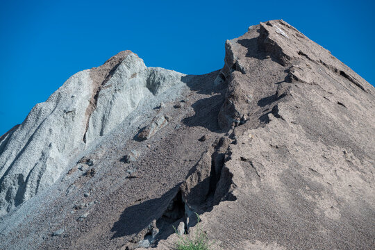 Towering mounds of excavated gravel and slag display jagged ridges and deep erosion channels set against a bright, clear blue sky.