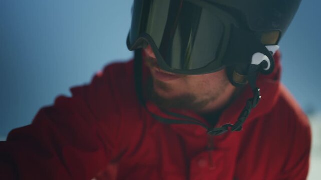 A male athlete wearing a red ski jacket, helmet, and goggles prepares for winter mountain sports. He gets ready to hit the snowy slopes on a bright, cold day.