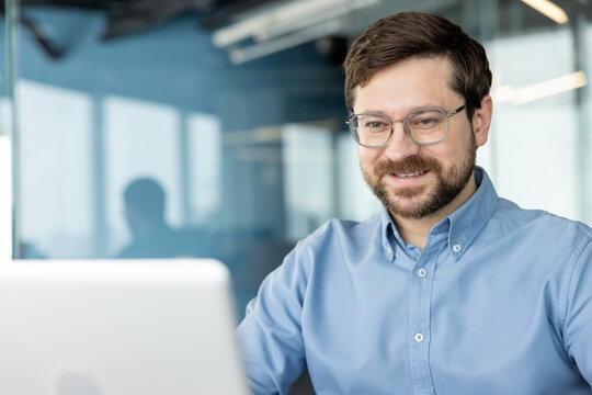A close-up photo of a young, smiling man wearing glasses, sitting at his workspace and working on a laptop