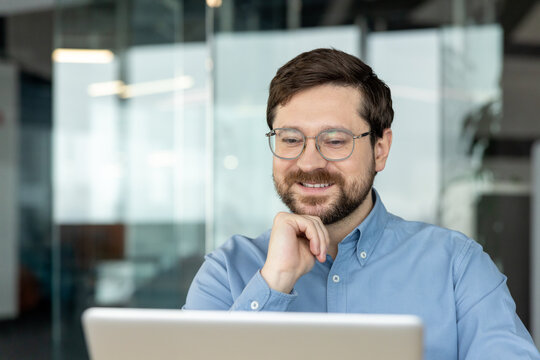A close-up photo of a young, smiling man wearing glasses, sitting at his workspace, holding his beard, and looking thoughtfully at a laptop screen
