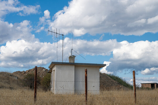A small, utilitarian white concrete building with rooftop television and radio antennas stands behind a barbed wire fence in an arid rural field under a vibrant cloudy sky.