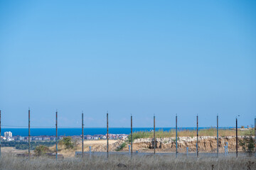 Fototapeta premium A wire fence lines a grassy ridge in the foreground, overlooking a distant coastal town and the deep blue sea under an expansive clear blue sky.