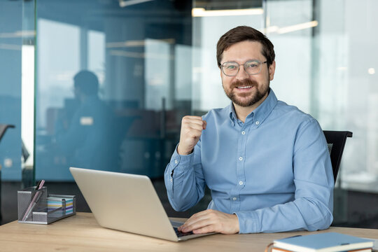 A portrait of a happy young man wearing glasses, sitting at an office desk with a laptop, looking at the camera, and making a victory sign with his hand