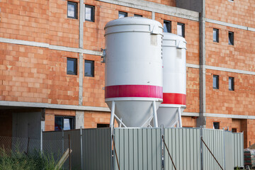 Two large industrial cement silos stand behind a metal fence at an active residential building construction site featuring an unplastered red clay brick facade. © Andrey