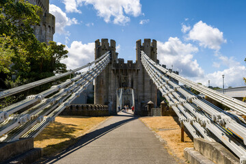 Fototapeta premium Conwy Suspension Bridge cables leading toward stone gatehouse, Conwy Castle towers behind under clear blue sky with white clouds, Wales