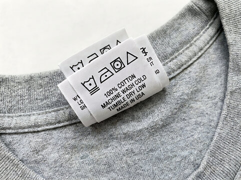 Close-up of a gray t-shirt with a care label displaying washing instructions and fabric composition, showing symbols for machine wash, tumble dry, and ironing