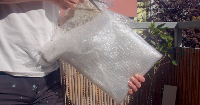 close-up hands peel transparent bubble wrap from honeycomb wooden board on sunny balcony, revealing cell pattern and warm light reflections in daylight glints