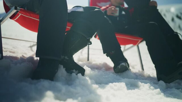 Individuals are seen relaxing in red chairs on a bright, snowy mountain landscape. Boots and snow are prominent in this winter leisure scene.