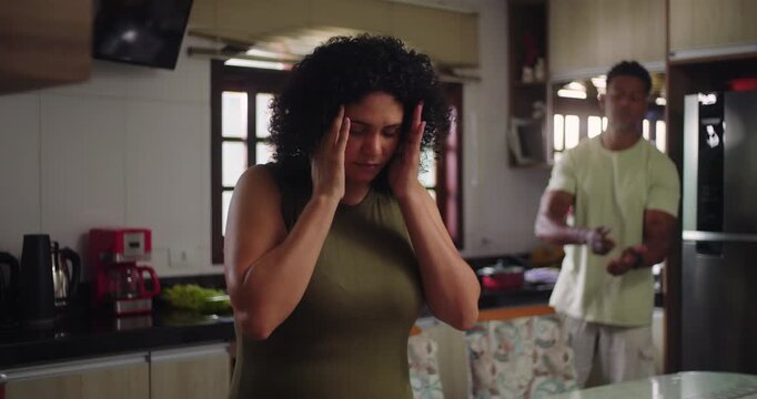 Wife visibly distressed with hands on head in foreground as husband gestures during argument in kitchen, showcasing emotional tension and strained relationship dynamics