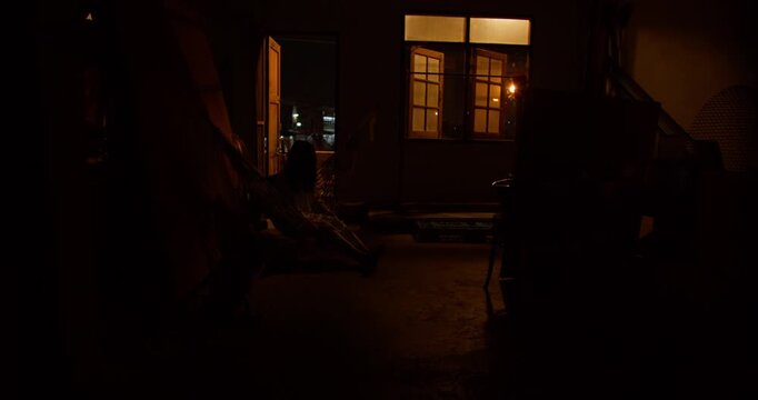 A ghostly figure sits motionless in a hammock within a dark and cluttered room, creating a scary and suspenseful atmosphere perfect for a horror film scene.