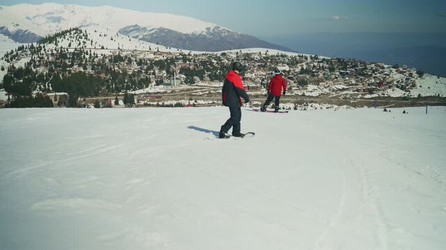 Two snowboarders glide down a pristine, snow-covered mountain slope. A beautiful ski resort and distant peaks complete the stunning winter landscape.