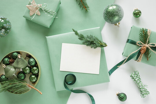 Overhead view of a blank card, wrapped Christmas gifts, a box of festive Christmas baubles, twine and assorted foliage on a white background