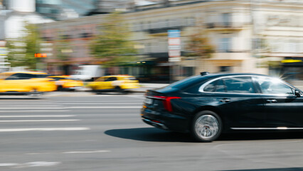 A luxury black sedan speeds past a line of yellow taxis in a bustling city. The scene captures the movement of vehicles against a backdrop of buildings and traffic signals © Sergio